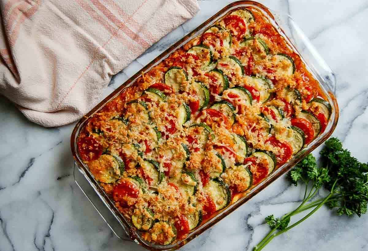 A baked zucchini casserole sits on a marble countertop. Sliced zucchini and tomatoes, are arranged in rows, topped with melted cheese and breadcrumbs. A beige towel and fresh parsley are nearby.