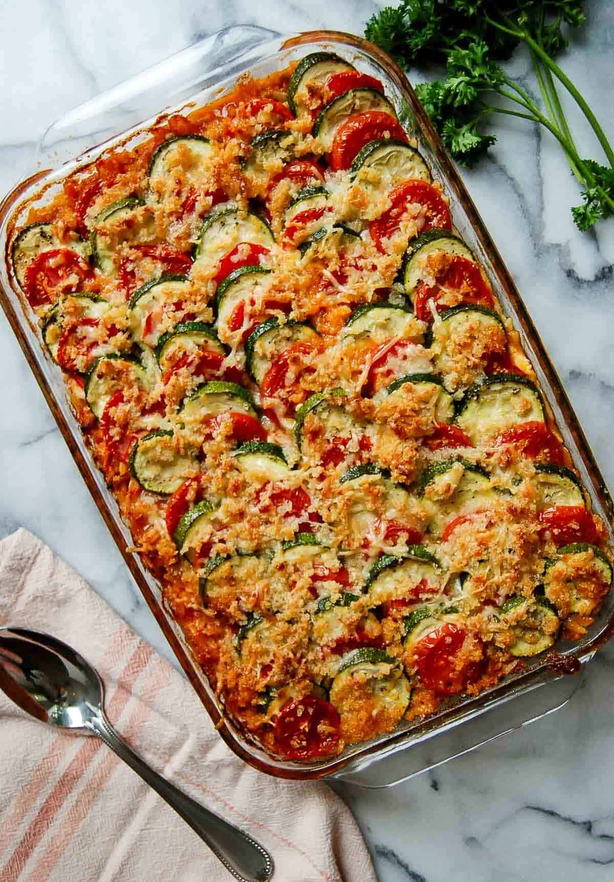 A glass baking dish filled with a baked vegetable gratin, layered with sliced zucchini and tomatoes, topped with a golden breadcrumb and cheese crust. A spoon, towel, and fresh parsley are nearby on a marble surface.