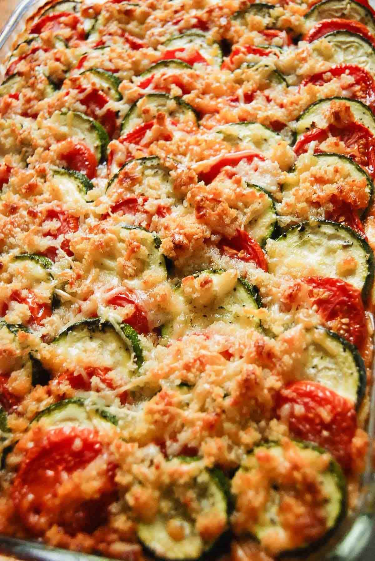 A close-up of a baked vegetable casserole featuring overlapping slices of zucchini and tomato, topped with melted cheese and golden-brown breadcrumbs.