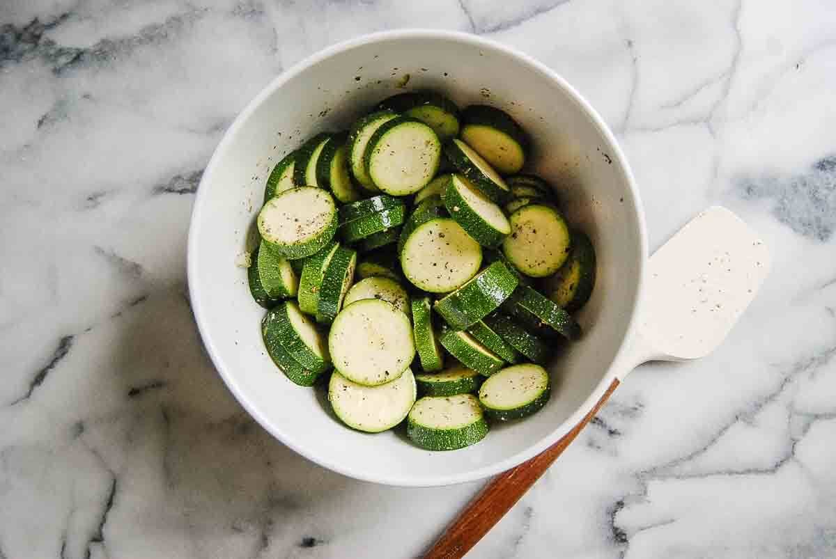 A white bowl filled with sliced zucchini rounds seasoned with black pepper, placed on a marble countertop next to a white spatula with a wooden handle.