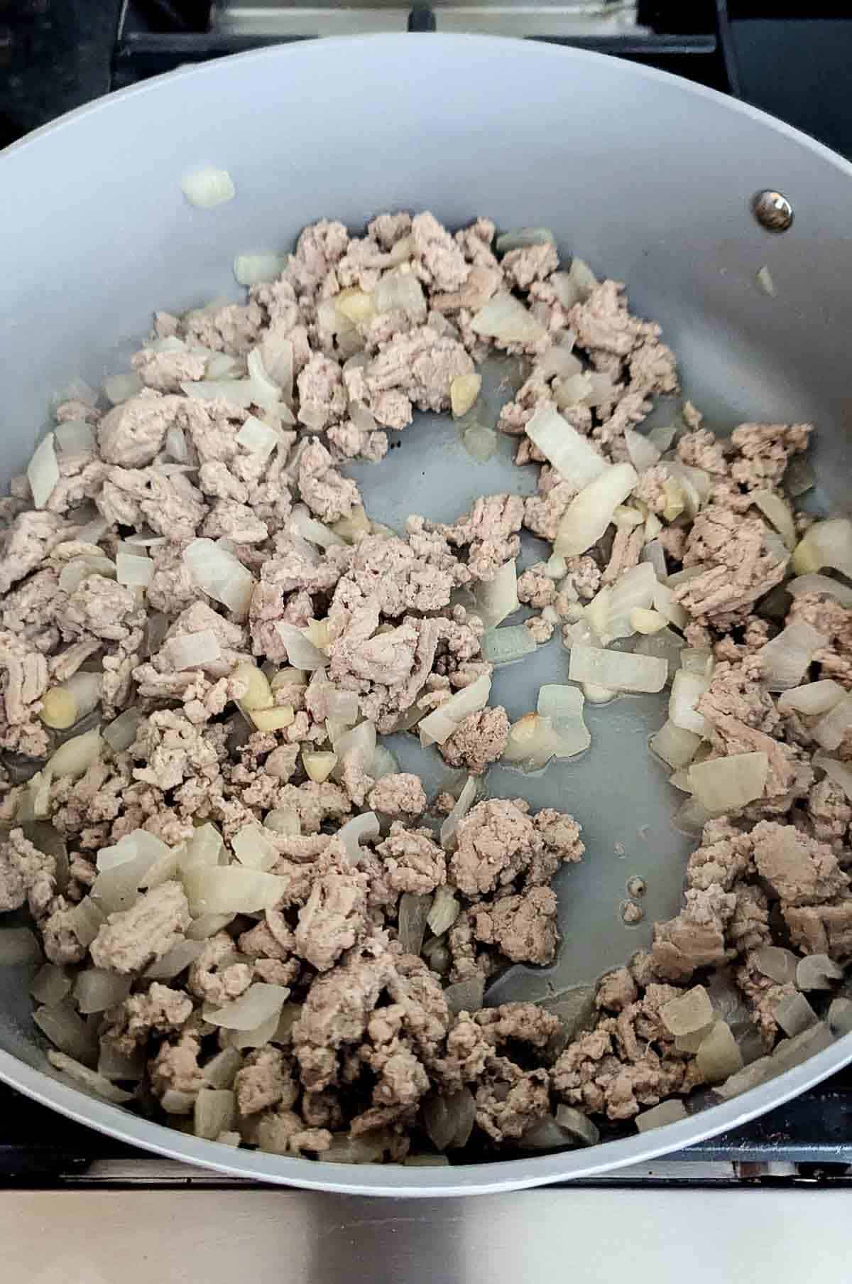 Ground turkey and chopped onions are being cooked in a large gray skillet on a stovetop. The mixture is browning and appears partially cooked.