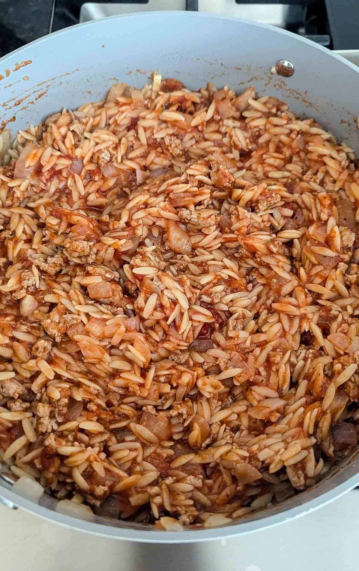 A large pan filled with cooked orzo pasta mixed with ground meat, chopped onions, and tomato sauce, creating a hearty and savory dish.