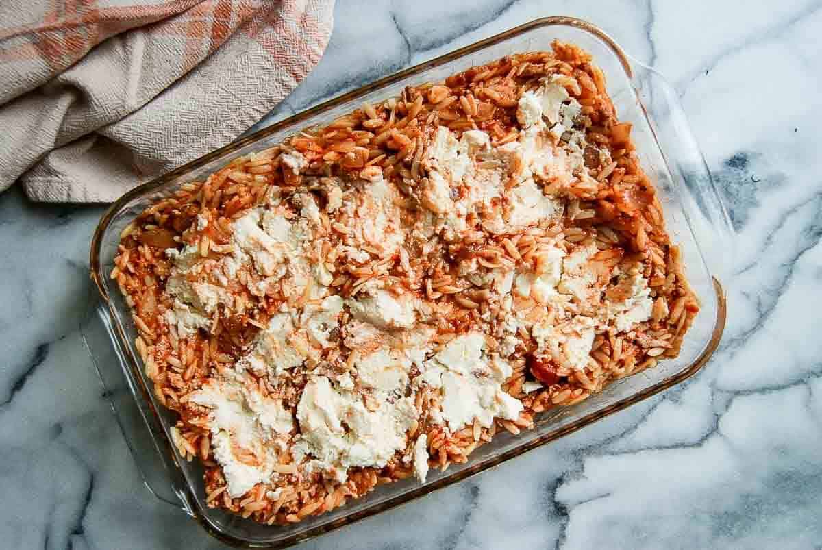 A glass baking dish filled with baked orzo pasta in tomato and ground turkey sauce, topped with dollops of ricotta cheese, sits on a marble surface next to a beige and pink checkered kitchen towel.