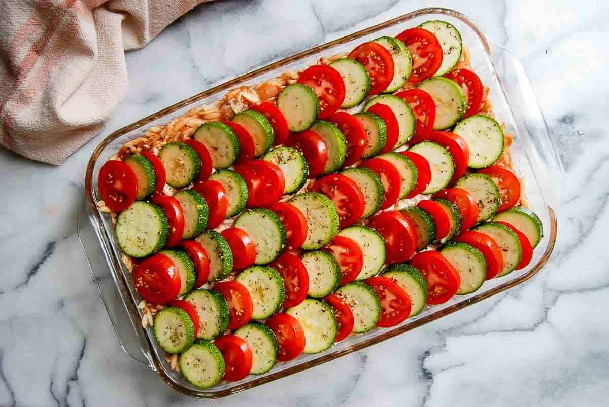 A glass baking dish filled with rows of sliced zucchini and tomatoes arranged neatly on top of a layer of sauce and cheese, ready to be baked, on a marble countertop.