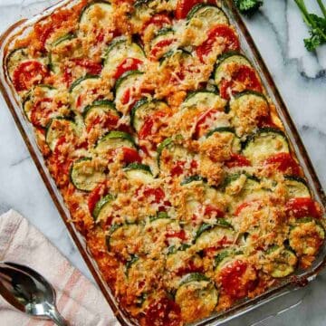A glass baking dish filled with a colorful baked vegetable casserole, layered with sliced zucchini, tomatoes, and topped with a golden, crispy breadcrumb and cheese topping. A serving spoon and napkin are nearby.