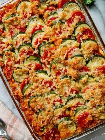 A glass baking dish filled with a colorful baked vegetable casserole, layered with sliced zucchini, tomatoes, and topped with a golden, crispy breadcrumb and cheese topping. A serving spoon and napkin are nearby.