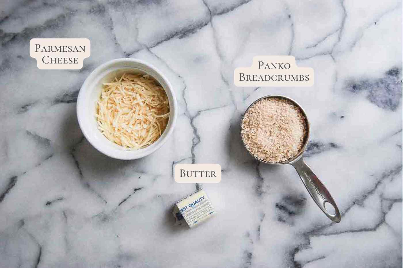 A bowl of shredded Parmesan cheese, a measuring cup of panko breadcrumbs, and a small stick of butter are arranged on a marble surface, each labeled with their names.