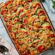 A glass baking dish filled with a baked vegetable casserole featuring sliced zucchini and tomatoes, topped with a golden, crispy breadcrumb and cheese topping. A spoon and striped towel are nearby, with fresh herbs in the corner.