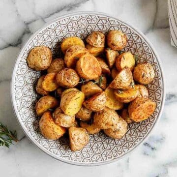 A patterned bowl filled with golden-brown roasted potato halves and quarters, garnished with herbs, sits on a marble surface.