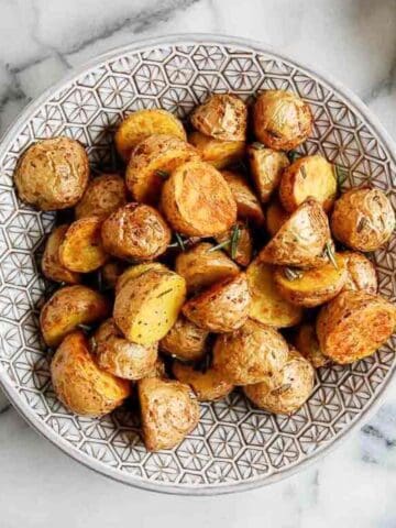A patterned bowl filled with golden-brown roasted potato halves and quarters, garnished with herbs, sits on a marble surface.