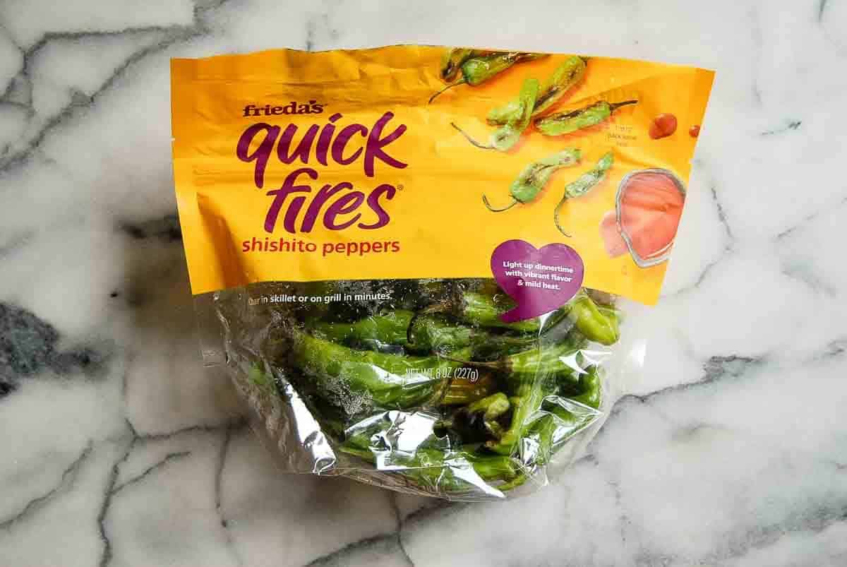 A package of Frieda’s Quick Fires shishito peppers sits on a marble surface. The top half of the bag is yellow-orange with images of peppers and a dipping sauce, while the bottom half is clear showing green peppers inside.