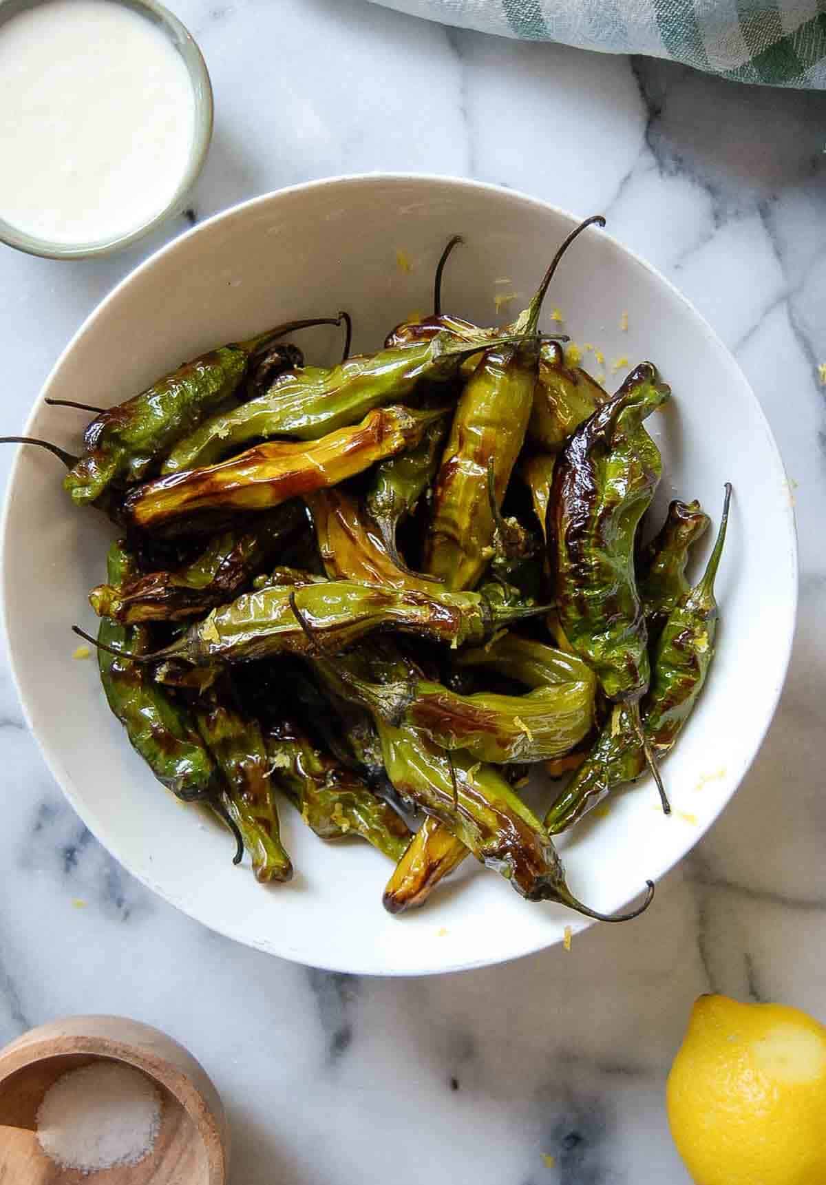A white bowl filled with blistered shishito peppers on a marble surface, with a lemon, a small wooden bowl of salt, and a cup of creamy dipping sauce nearby.