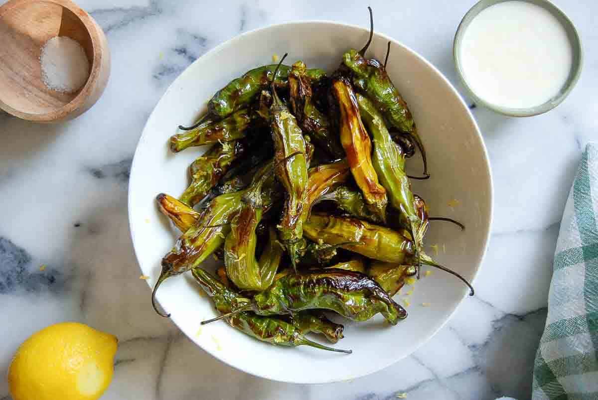 A bowl of blistered shishito peppers on a marble surface, with a small wooden bowl of salt, a lemon, a cup of dipping sauce, and a green checked napkin nearby.