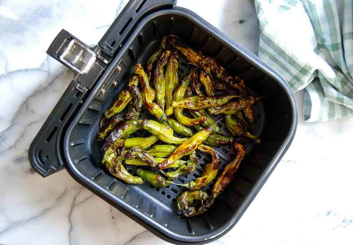 Charred shishito peppers in an open air fryer basket on a marble countertop, with a green and white striped kitchen towel nearby.