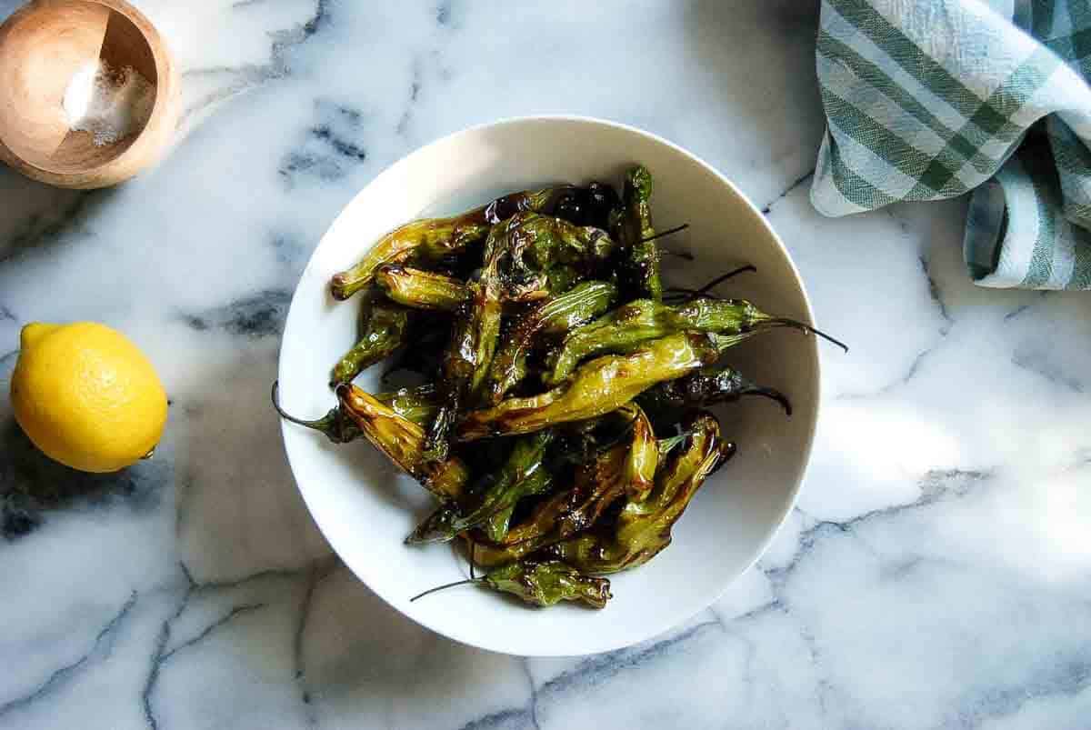A white bowl filled with blistered shishito peppers sits on a marble surface next to a lemon, a small wooden bowl of salt, and a green-and-white striped cloth.