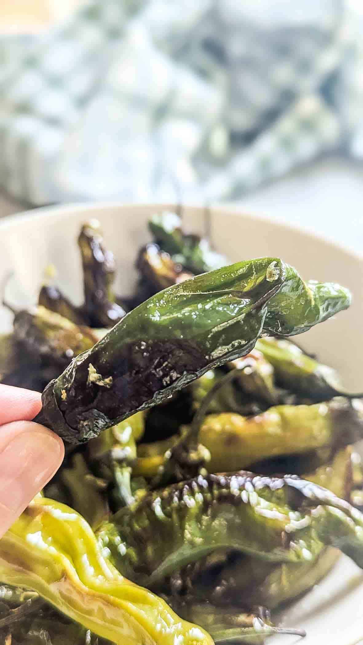 A hand holds a blistered, roasted shishito pepper above a bowl filled with more roasted peppers; the background is softly blurred.