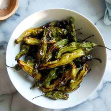 A white bowl filled with blistered, roasted shishito peppers sits on a marble surface. The peppers are charred in spots and vary in shades of green and yellow.