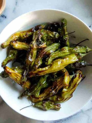 A white bowl filled with blistered, roasted shishito peppers sits on a marble surface. The peppers are charred in spots and vary in shades of green and yellow.