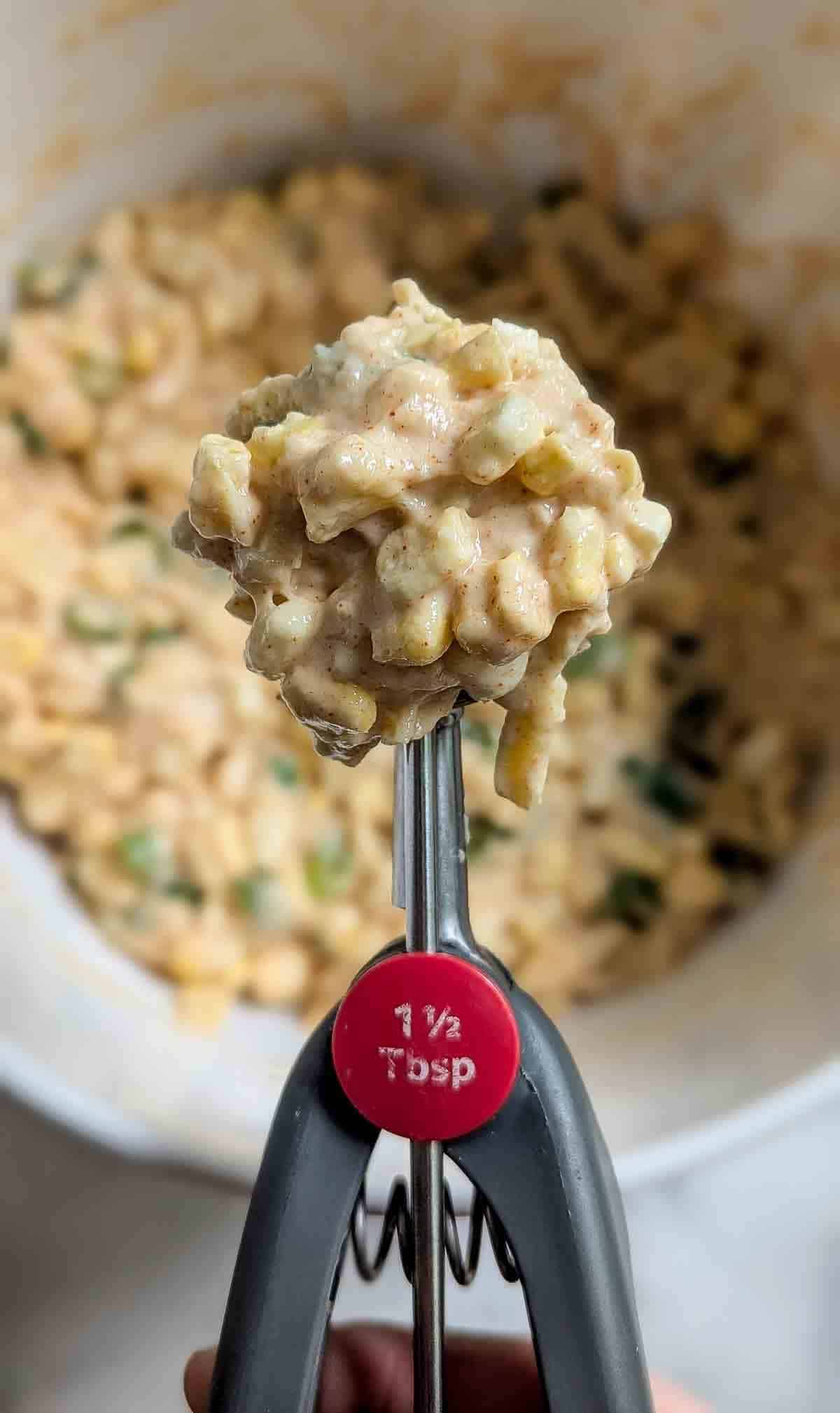 A close-up of a cookie scoop labeled 1 ½ tablespoon holding a portion of thick, chunky batter over a bowl filled with a similar mixture in the background.