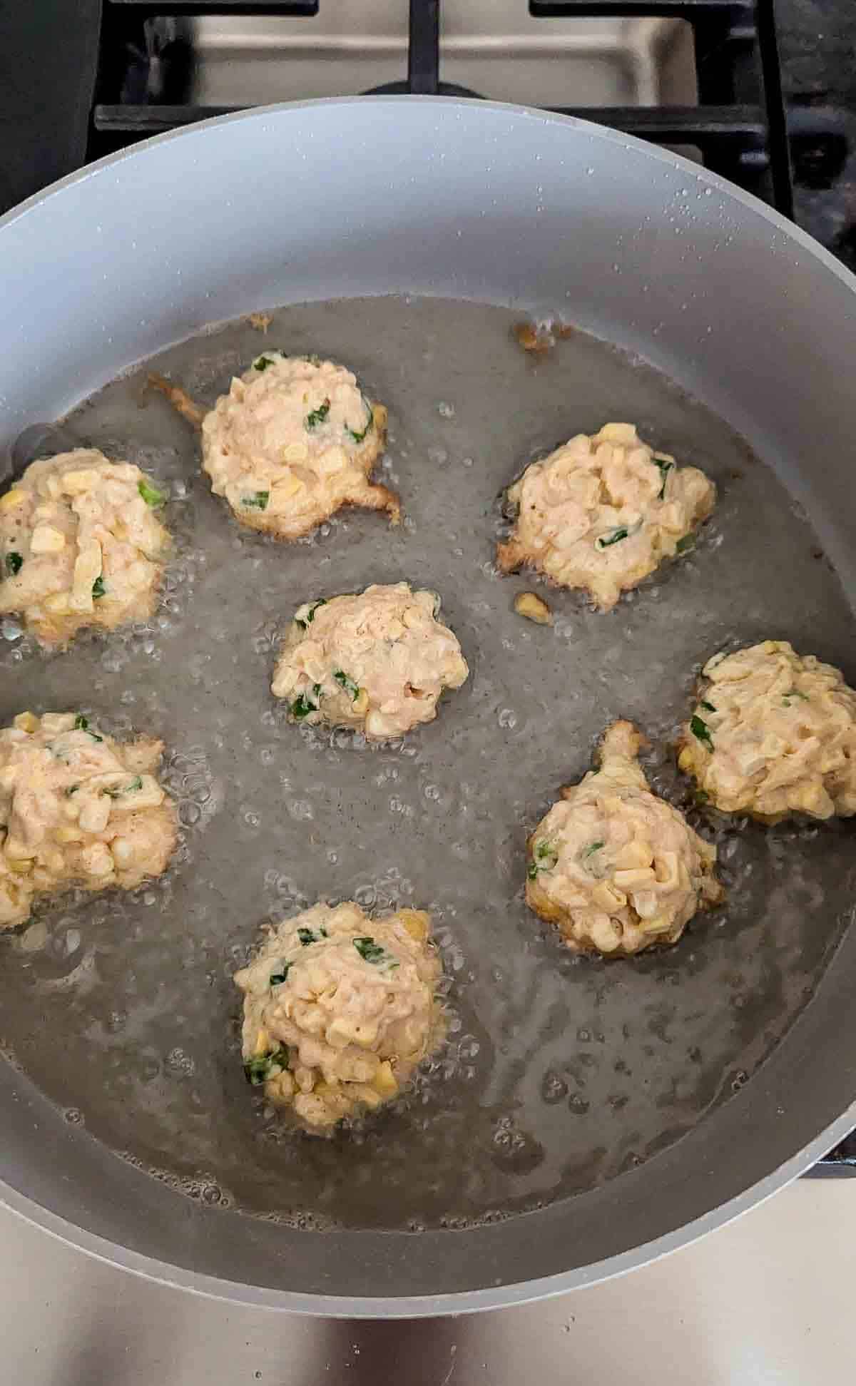 Seven fritters made of a light batter with visible corn kernels and herbs are being deep-fried in bubbling oil in a grey pan on a stovetop.