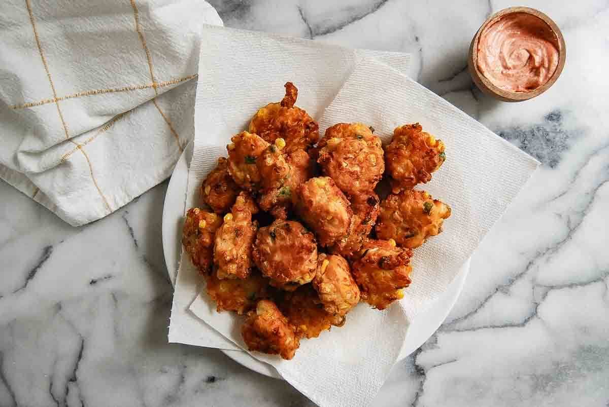 A plate lined with paper towels holds a pile of golden-brown fritters, with a dish of creamy dipping sauce and a folded white towel nearby on a marble surface.