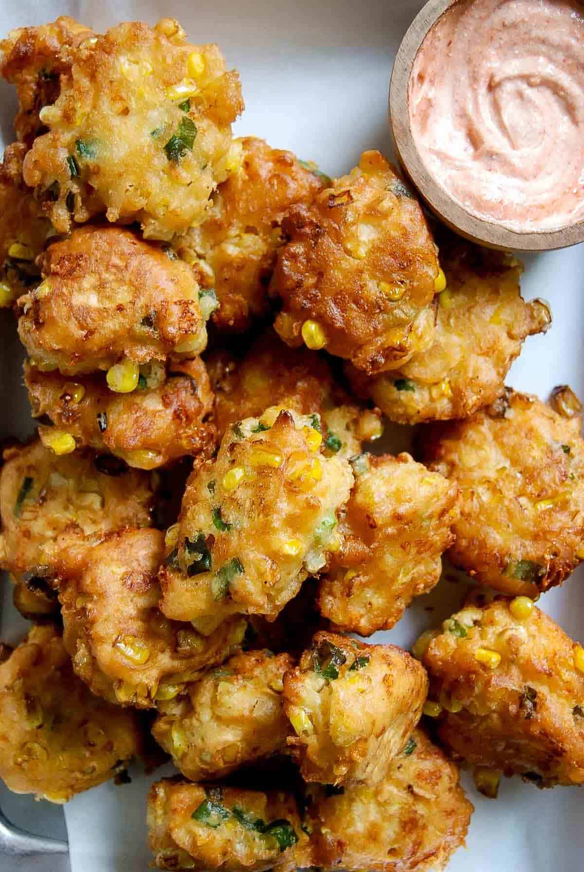 A plate of golden-brown corn fritters mixed with herbs, served with a small wooden bowl of creamy dipping sauce.