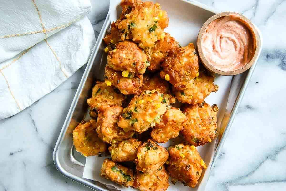 A tray of golden-brown corn fritters garnished with herbs, served with a small bowl of creamy dipping sauce on the side, sits on a marble surface next to a folded white cloth.