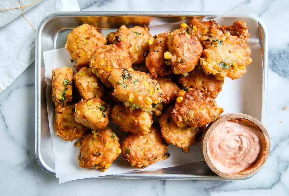 A tray of golden-brown fritters with corn and herbs, served on parchment paper with a small bowl of creamy dipping sauce on the side.