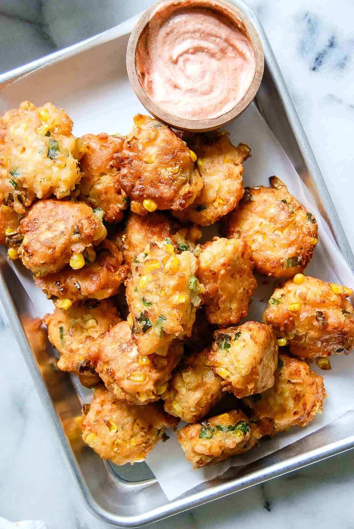 A metal tray filled with golden-brown corn fritters garnished with herbs, served with a small bowl of creamy dipping sauce on the side. The tray sits on a marble surface.