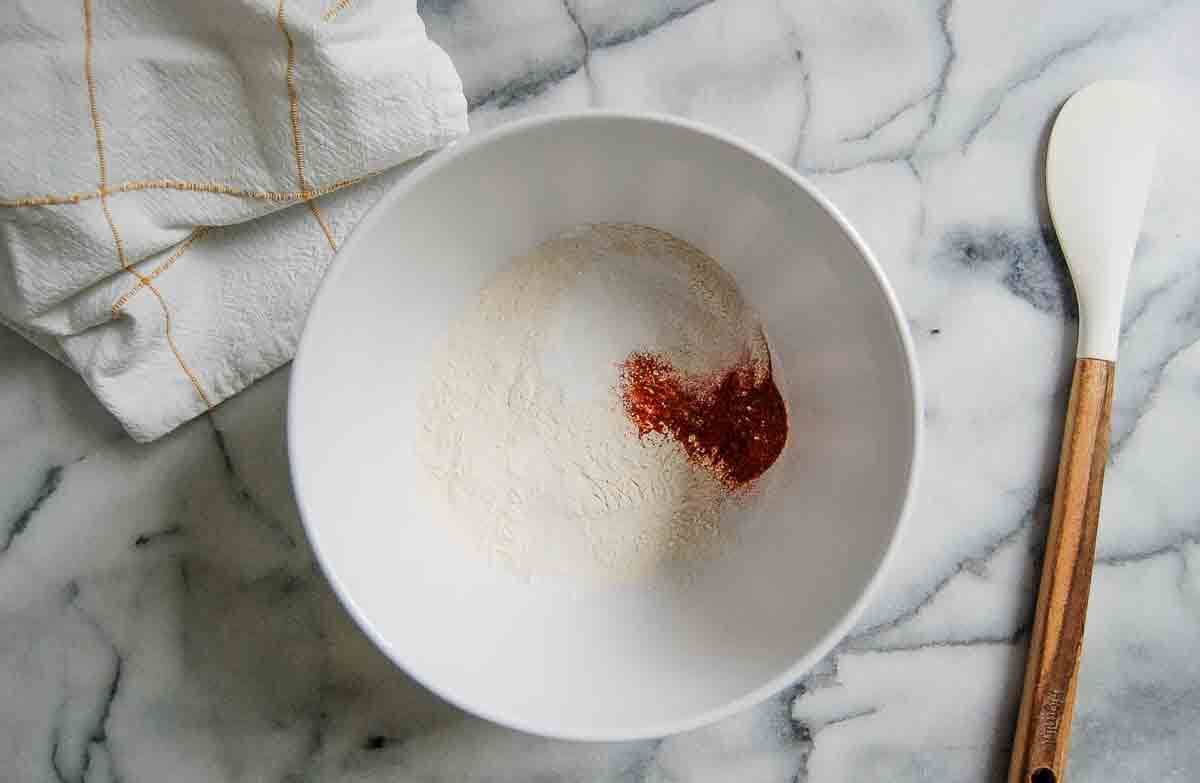 A white mixing bowl with flour and a reddish spice sits on a marble countertop next to a white spatula with a wooden handle and a folded white dish towel with yellow lines.