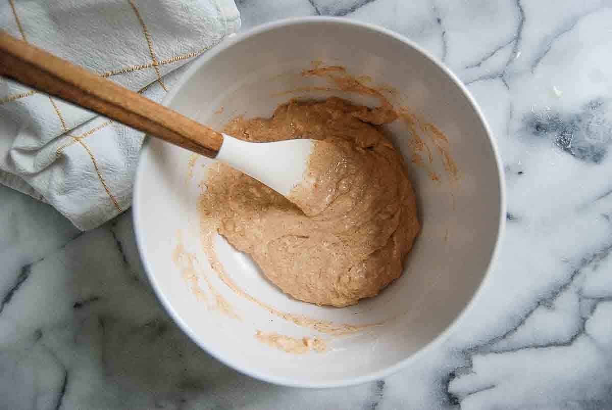 A white mixing bowl sits on a marble countertop, holding thick, beige batter. A spatula with a wooden handle rests in the bowl. A white kitchen towel with yellow stripes is nearby.