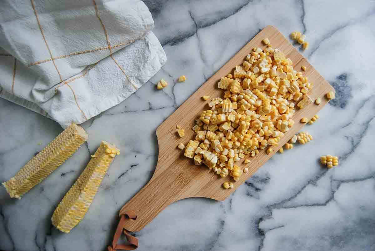 Chopped corn kernels on a wooden cutting board, with two empty corn cobs and a white kitchen towel on a marble countertop.