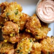 Golden corn fritters with visible corn kernels and green herbs are stacked on a plate next to a small bowl of creamy, pinkish dipping sauce.