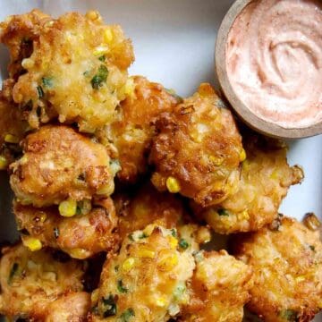 Golden corn fritters with visible corn kernels and green herbs are stacked on a plate next to a small bowl of creamy, pinkish dipping sauce.
