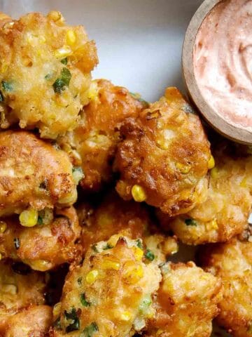 Golden corn fritters with visible corn kernels and green herbs are stacked on a plate next to a small bowl of creamy, pinkish dipping sauce.
