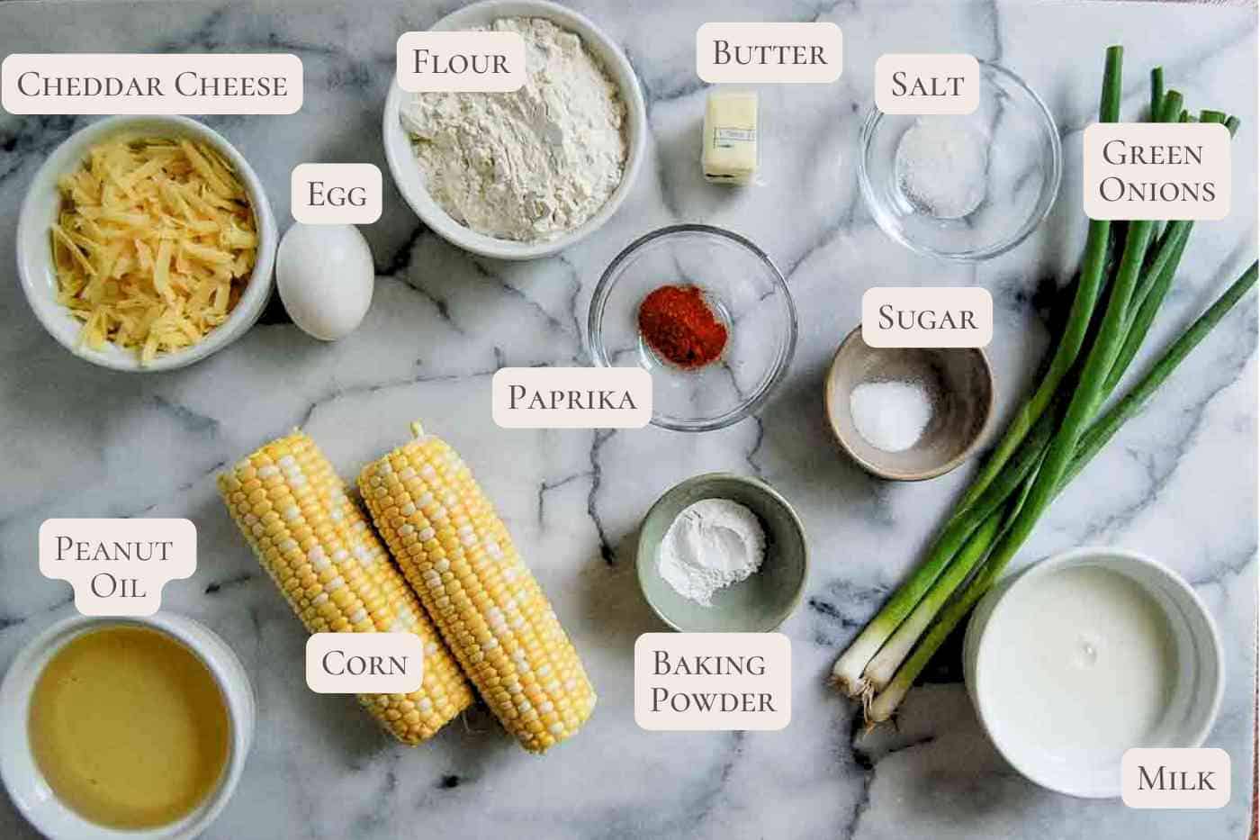 Various ingredients are arranged on a marble surface, including corn cobs, green onions, shredded cheddar cheese, an egg, flour, butter, salt, sugar, peanut oil, baking powder, paprika, and a bowl of milk.