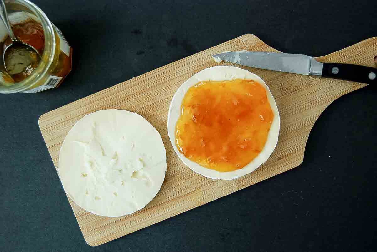 A round of soft cheese on a wooden board is sliced in half. One half is spread with apricot preserves. A jar of apricot preserves with a spoon and a knife are nearby on a dark surface.