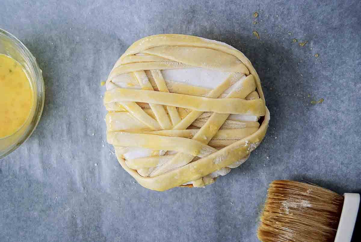 A round wheel of brie sits on parchment paper, wrapped in strips of puff pastry dough to resemble a mummy. An egg wash sits beside the wheel, with a pastry brush on the side.