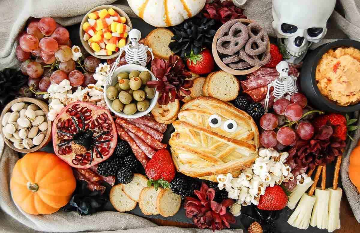 A festive Halloween charcuterie board with crackers, salami, fruit, olives, popcorn, dips, a mummy-shaped pastry, candy corn, pumpkins, a skull, plastic skeletons, and flowers, all arranged on a dark cloth.