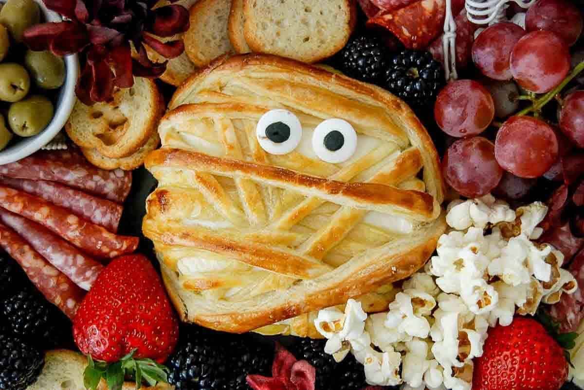 A round pastry wrapped in golden, crisscrossed dough to look like a mummy with two candy eyes, surrounded by grapes, strawberries, blackberries, olives, popcorn, crackers, and sliced cured meats.