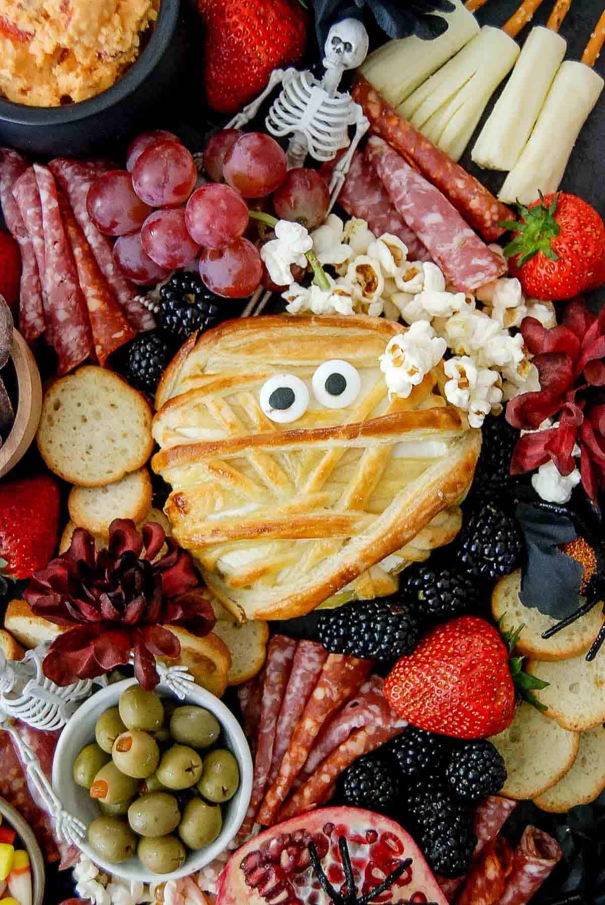 A Halloween charcuterie board with crackers, salami, grapes, olives, strawberries, popcorn, candy corn, chocolate pretzels, a pomegranate with a spider, and a mummy-shaped pastry with candy eyes.