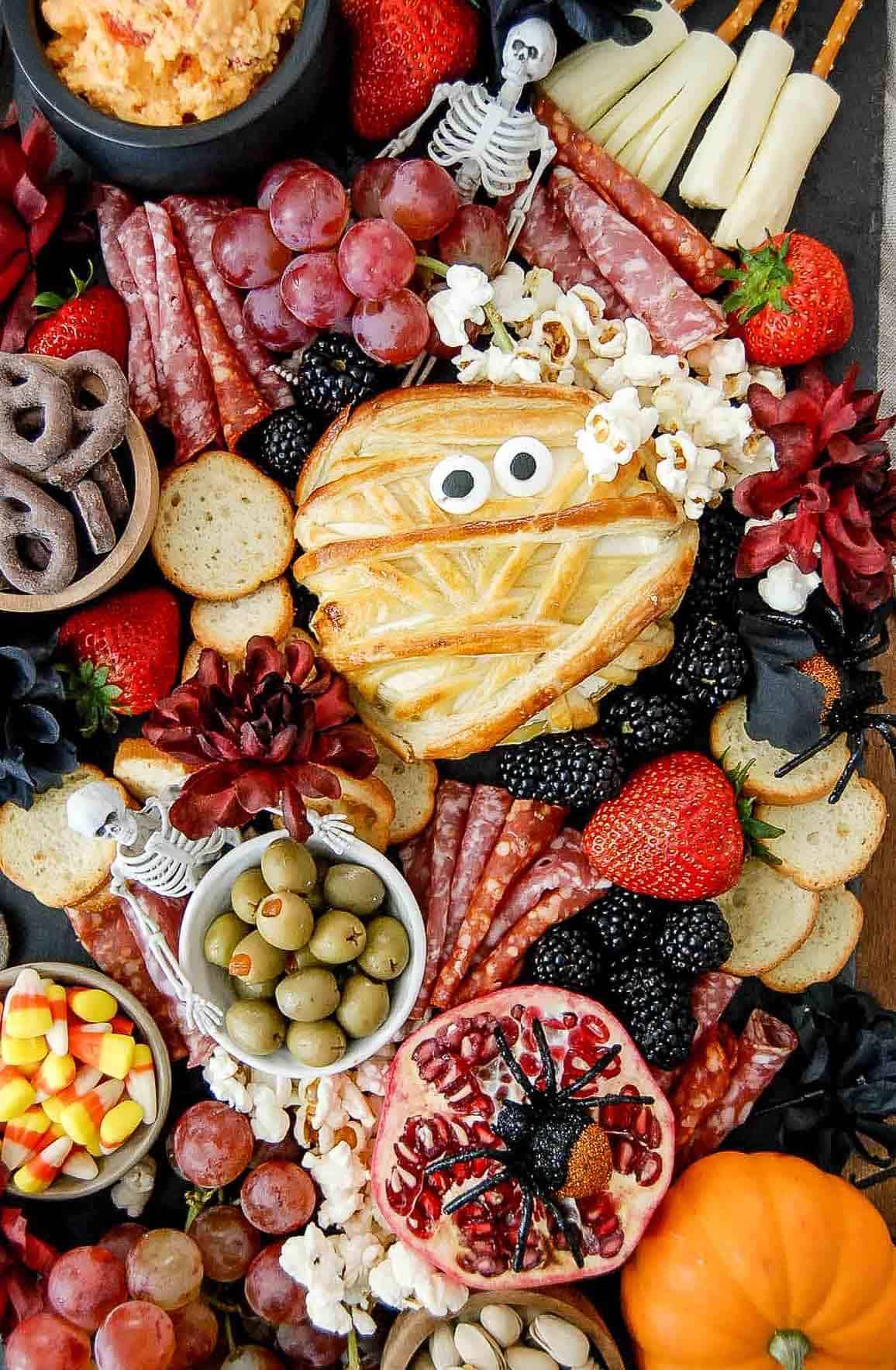 A Halloween charcuterie board with crackers, salami, grapes, olives, strawberries, popcorn, candy corn, chocolate pretzels, a pomegranate with a spider, and a mummy-shaped pastry with candy eyes.
