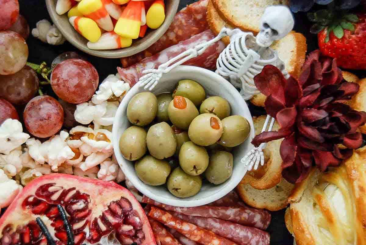 A festive charcuterie board with green olives, grapes, pomegranate, popcorn, candy corn, salami, toast, a strawberry, and a decorative plastic skeleton surrounded by flowers.