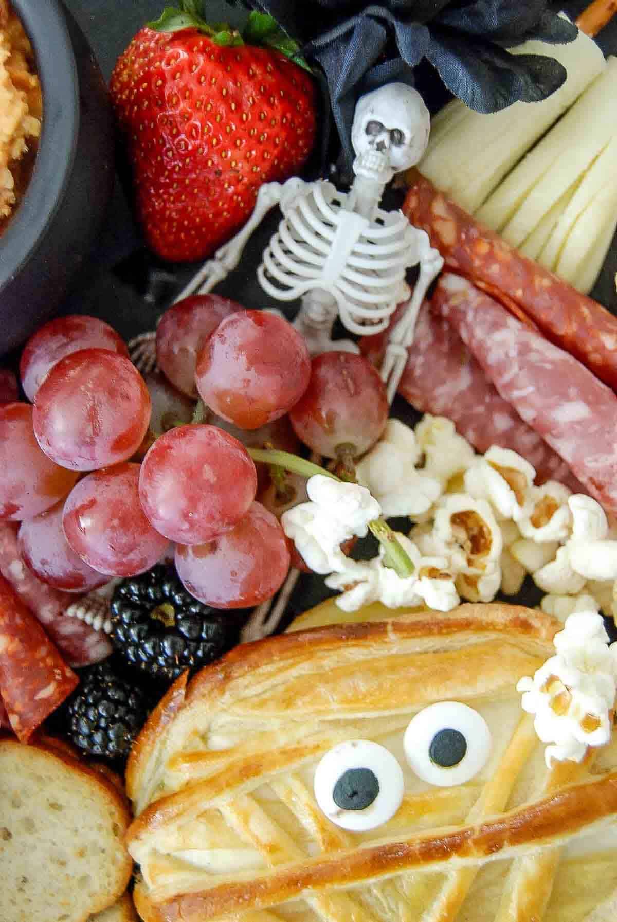 A Halloween-themed snack platter featuring a pastry decorated like a mummy with candy eyes, popcorn, grapes, cured meats, cheese slices, a strawberry, a blackberry, and a small plastic skeleton.