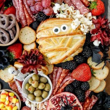 A Halloween charcuterie board with crackers, salami, grapes, olives, strawberries, popcorn, candy corn, chocolate pretzels, a pomegranate with a spider, and a mummy-shaped pastry with candy eyes.