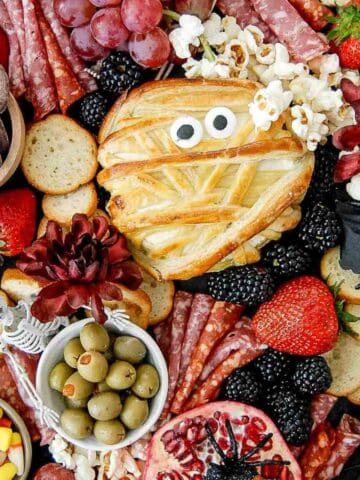 A Halloween charcuterie board with crackers, salami, grapes, olives, strawberries, popcorn, candy corn, chocolate pretzels, a pomegranate with a spider, and a mummy-shaped pastry with candy eyes.