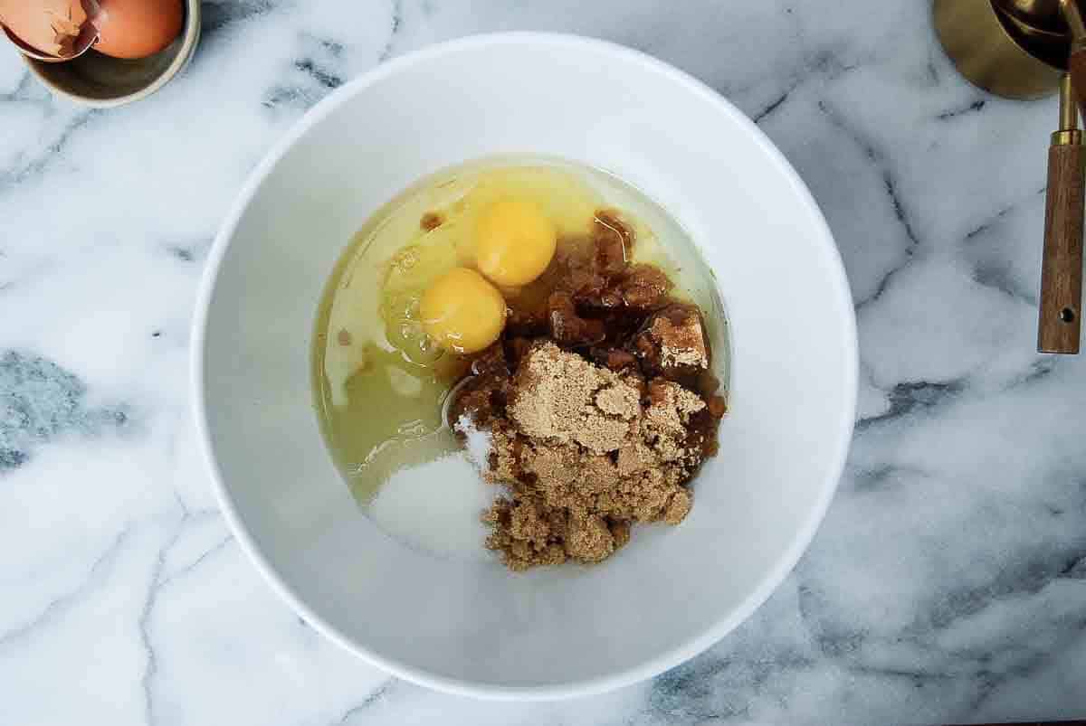 A white mixing bowl on a marble countertop contains two eggs, brown sugar, and white sugar, ready to be mixed. A small bowl of eggs and a measuring cup are nearby.
