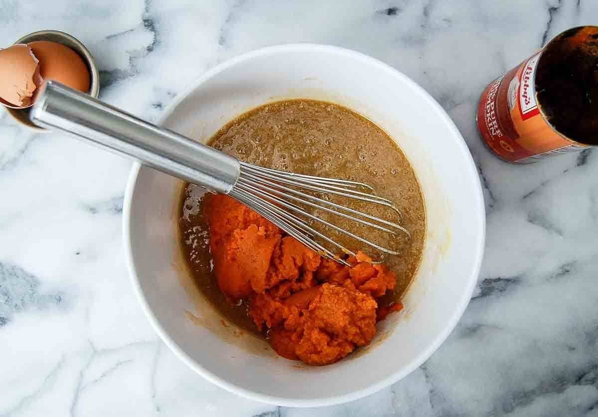 A whisk rests in a white bowl containing brown batter and orange pumpkin puree. An eggshell sits nearby, and a bottle of vanilla extract is partially visible on a marble countertop.