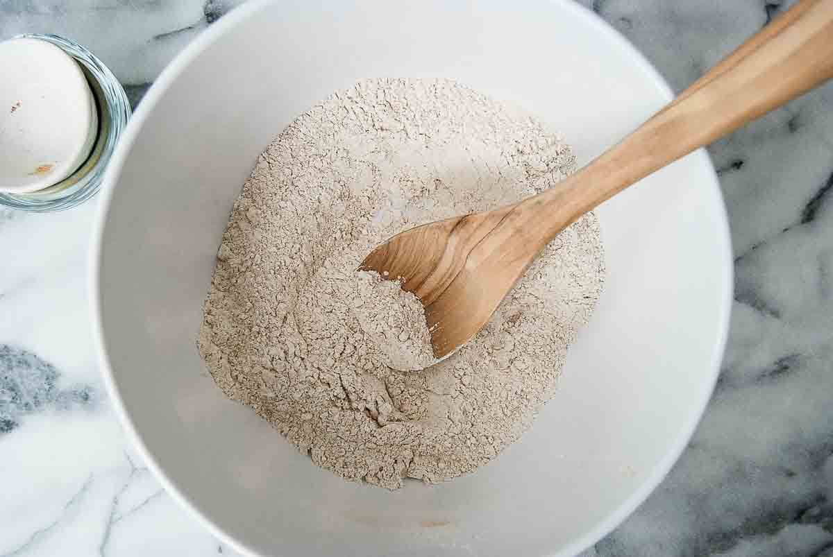 A wooden spoon rests in a white bowl filled with a mixture of dry ingredients on a marble countertop; an egg in a small glass container sits nearby.