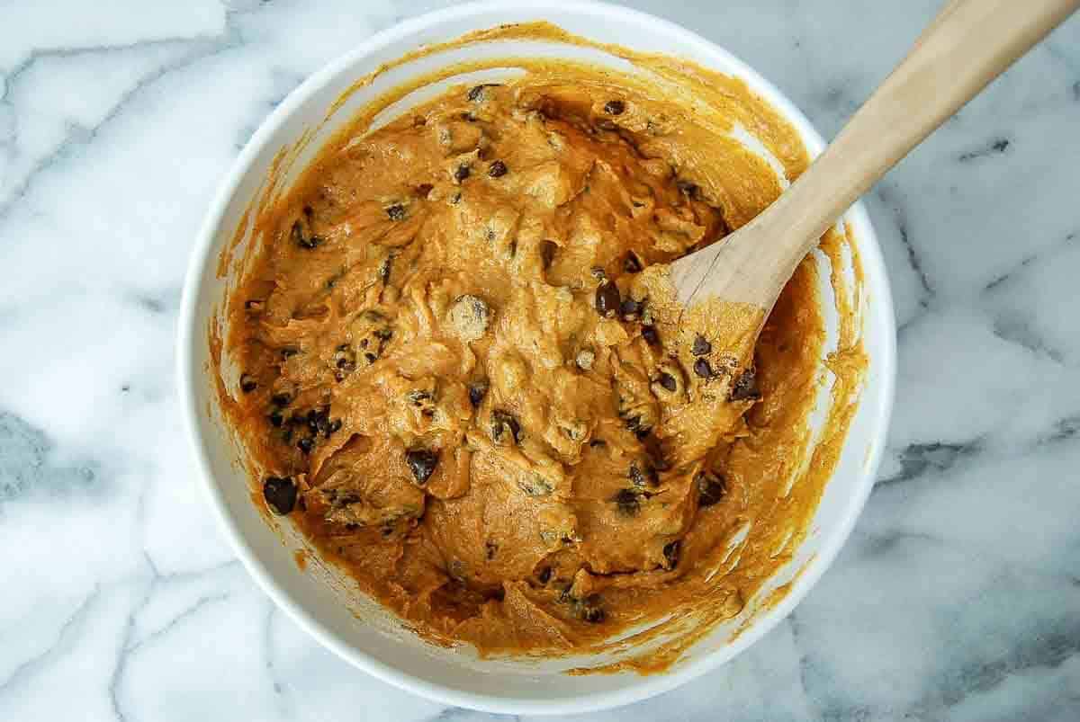 A white bowl filled with cookie dough mixed with chocolate chips sits on a marble surface. A wooden spoon is partially submerged in the dough, which has an orange-brown color.